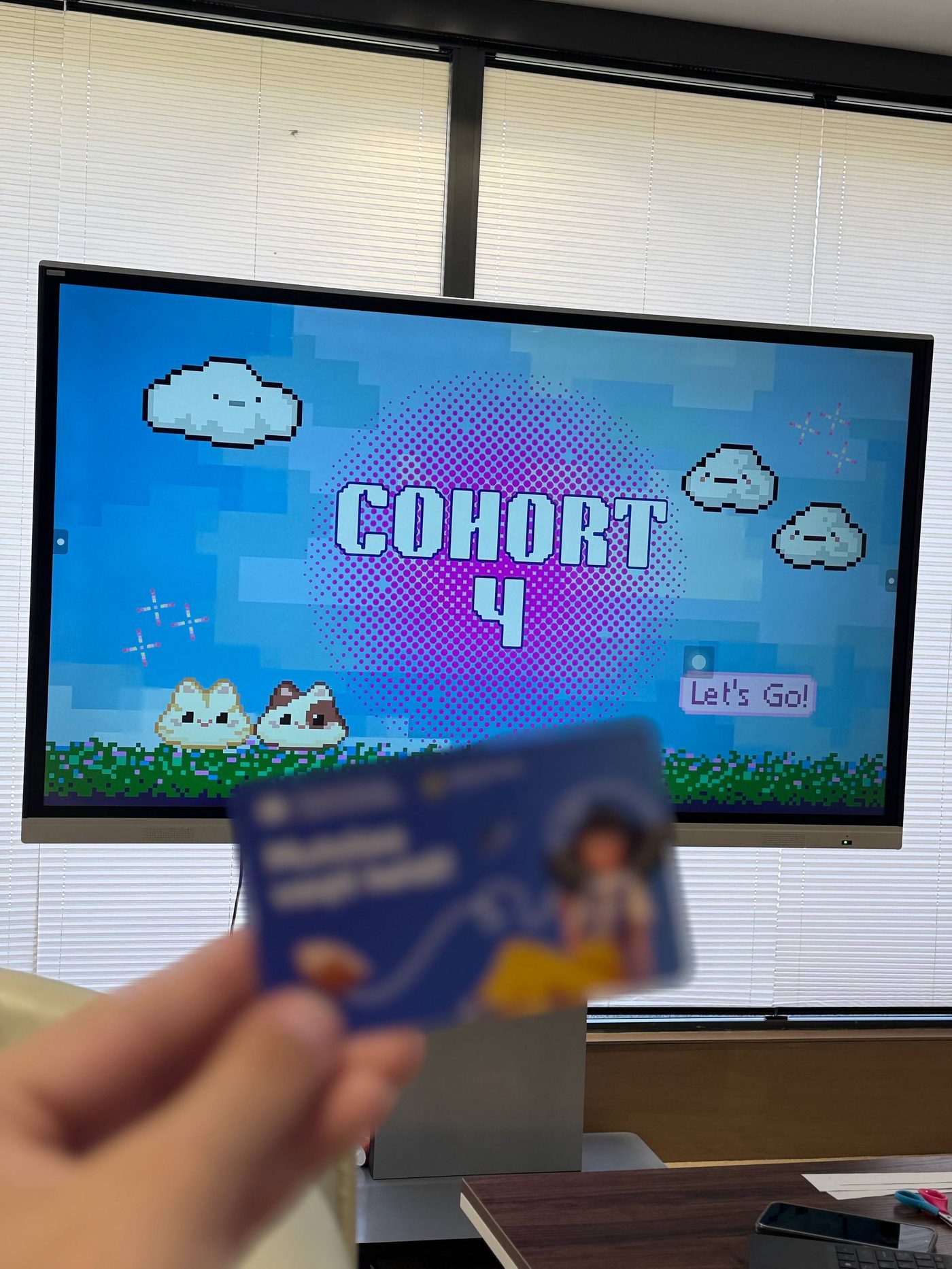 A screen displaying 'Cohort 4, Let's Go!' in a pixel-art style, with a participant's ID card in front